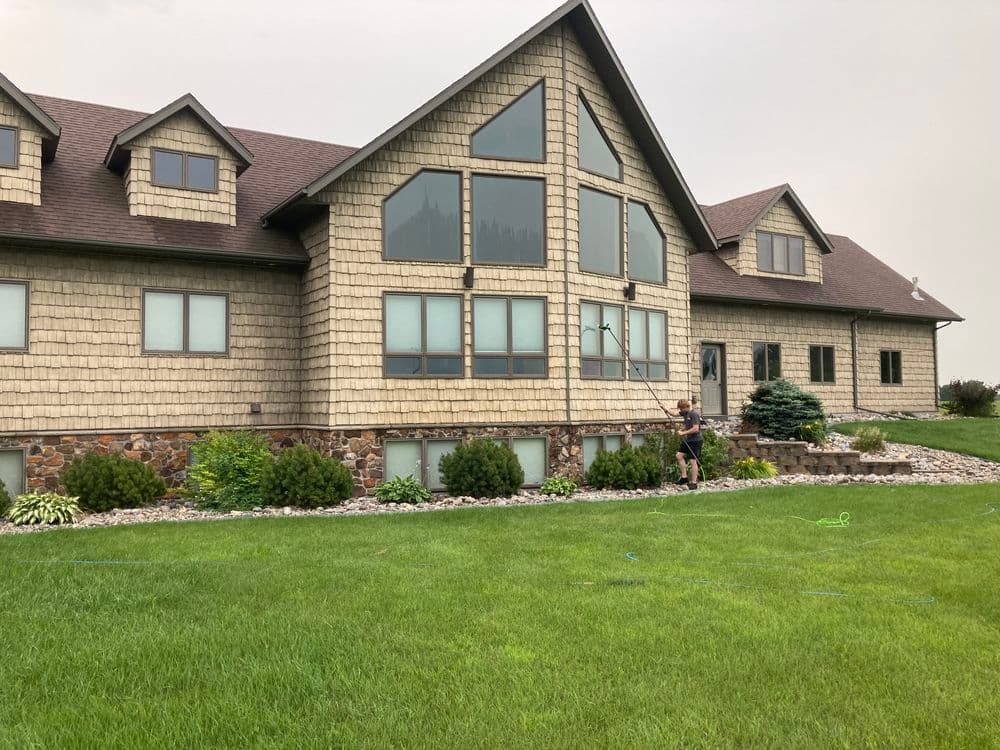 Professional window cleaner using a water-fed pole on a modern home with stone and wood siding.
