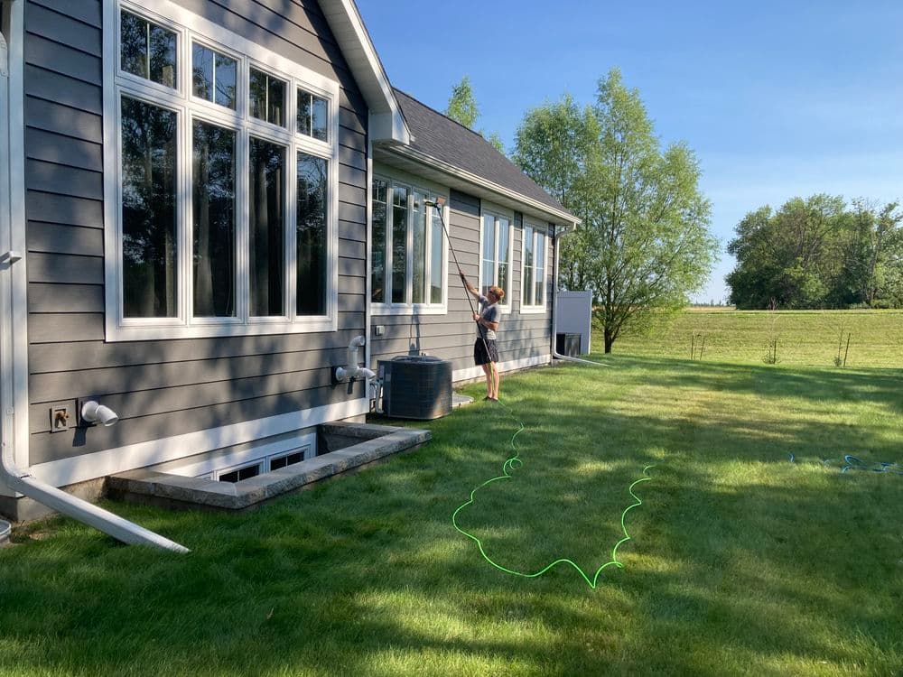 Person cleaning windows of a modern home with a ladder and green hose on a sunny day.