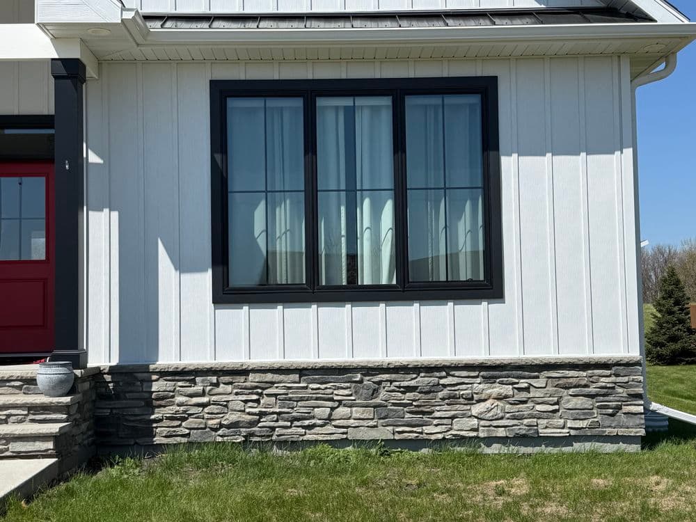 Modern home exterior featuring a large black-framed window and stone base on a sunny day.