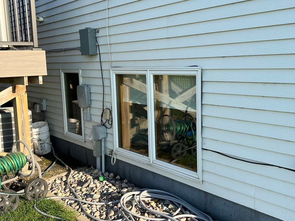 Side view of a house featuring two windows, utility connections, and a garden hose setup.