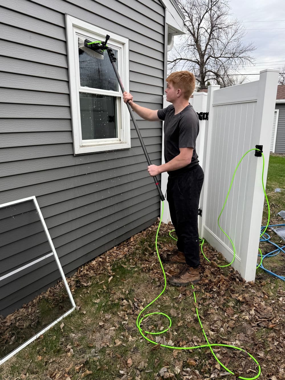 Young man using an extension pole to clean a window outside a house with gray siding.