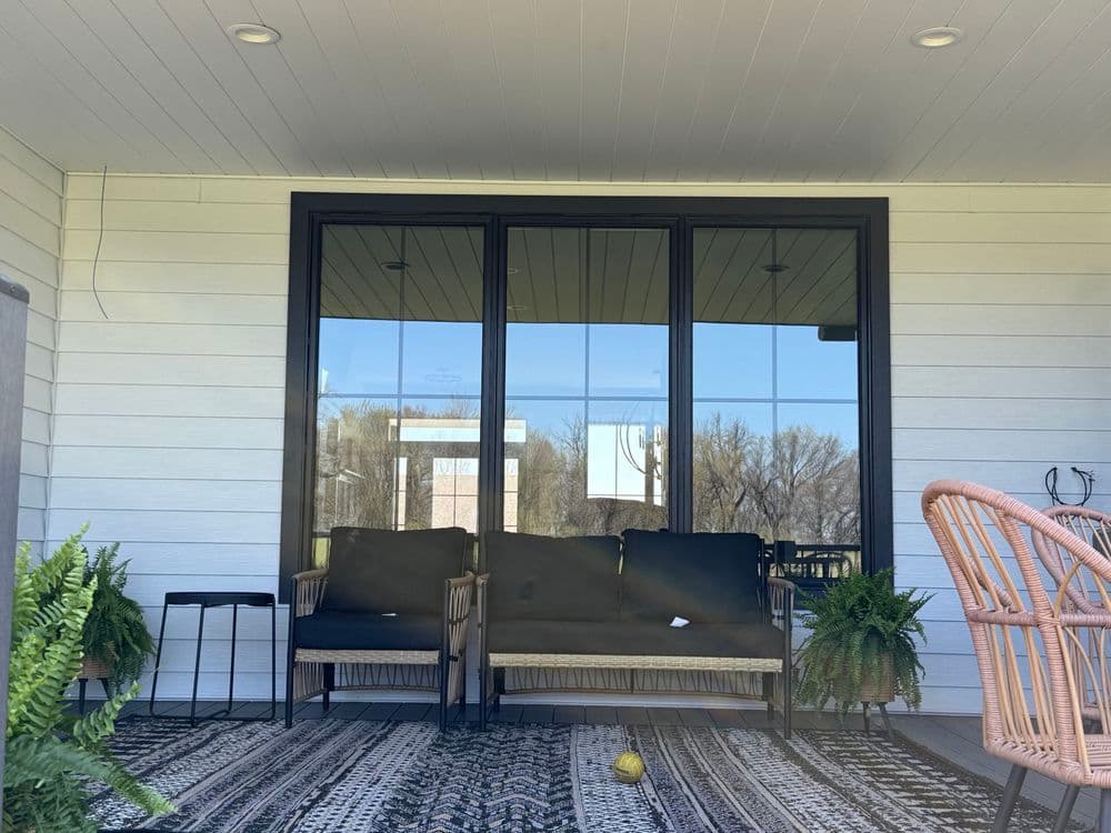 Cozy porch with black sofas, greenery, and a patterned rug near large windows.