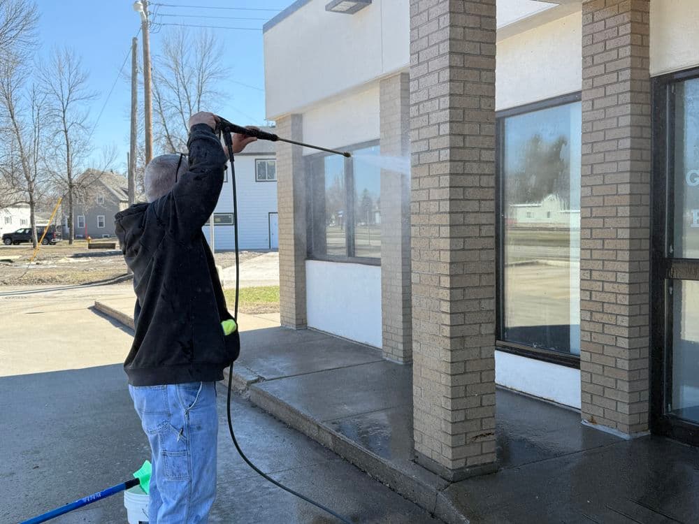 Man using a pressure washer to clean a building's exterior windows and entrance.