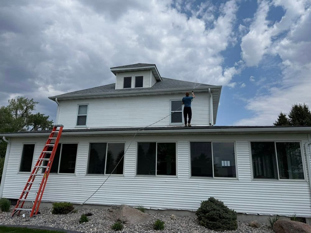 Worker using a paint roller on a house roof with a ladder under a cloudy sky.
