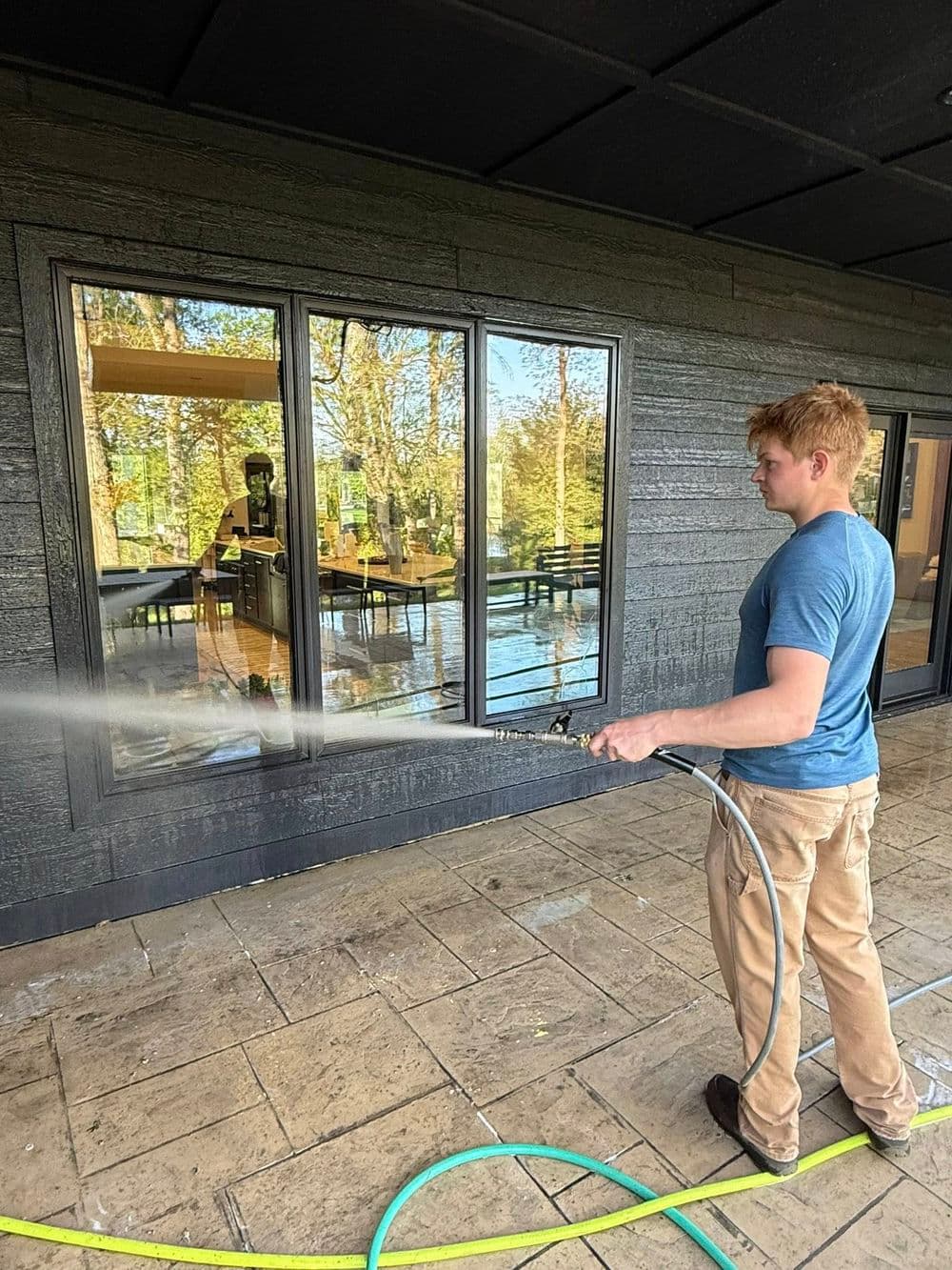 Man pressure washing large glass windows on a modern house exterior with trees in the background.