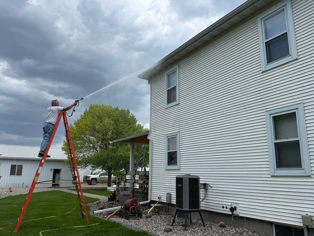 Person on ladder power washing a house exterior under a cloudy sky.
