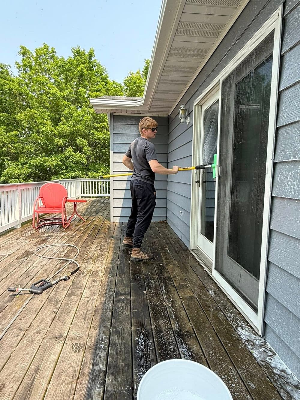 Man cleaning glass door on deck with squeegee, surrounded by trees and outdoor furniture.